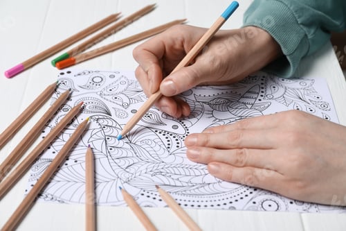 Preview: Woman coloring antistress picture with pencils at white table, closeup