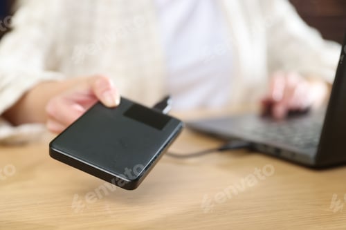 Preview: Woman connecting external hard drive to laptop at wooden desk, closeup