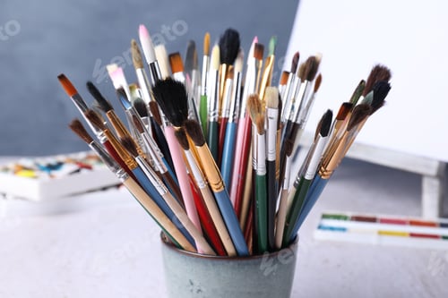 Preview: Paintbrushes, paints and easel with canvas on table near blue wall, closeup
