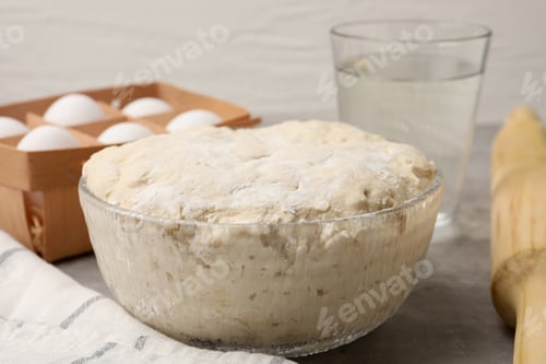 Preview: Fresh yeast dough in bowl, eggs, glass of water and rolling pin on gray table, closeup