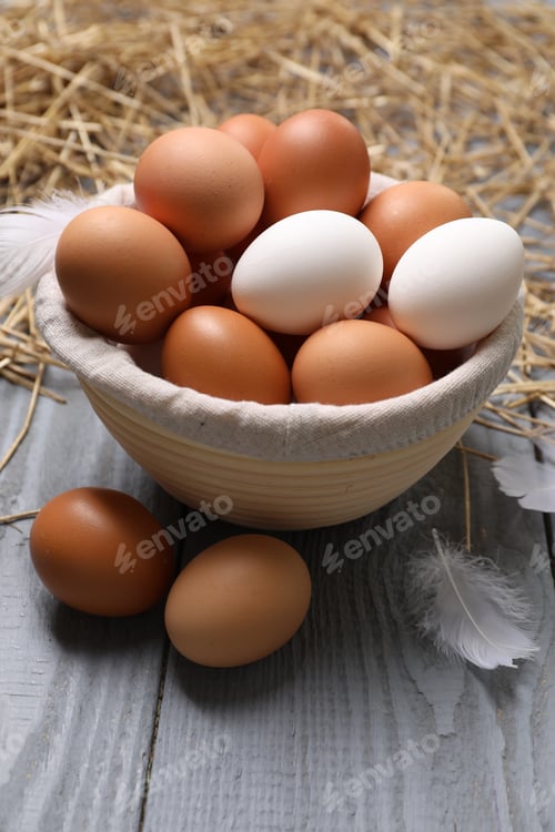 Preview: Fresh chicken eggs in bowl, feathers and dried straw on grey wooden table