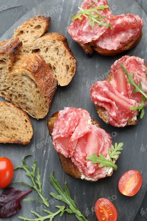 Preview: Delicious bruschettas with salami sausage, cream cheese, tomato and arugula on dark textured table,
