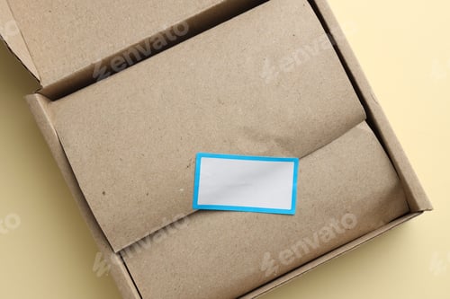 Preview: Cardboard box with packaging and blank sticker on beige background, top view