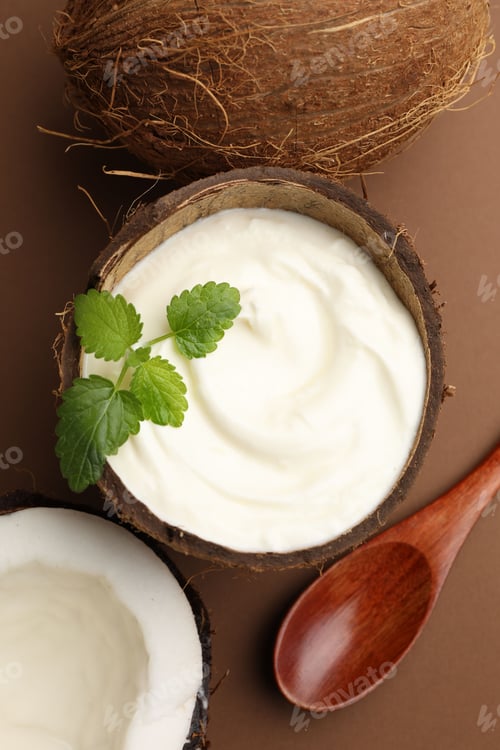 Preview: Tasty coconut yogurt with melissa leaves in shell and nuts on brown background, flat lay