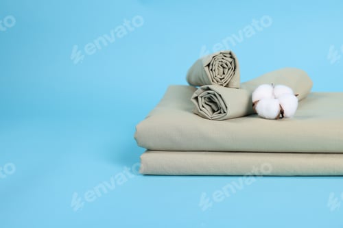 Preview: Stack of fresh bed linens and cotton flower on light blue background, closeup. Space for text