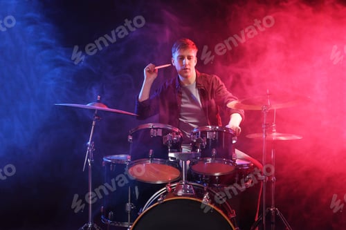 Preview: Man playing drums with drumsticks in color lights and smoke against black background