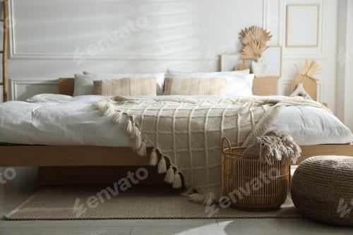 Vorschau: Großes Bett mit sauberer Bettwäsche, Weidenkorb und Hocker im Schlafzimmer. Innenarchitektur