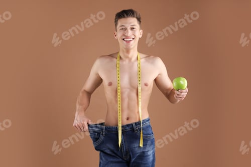 Preview: Athletic man with measuring tape and green apple wearing big jeans on brown background