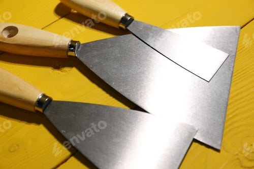 Preview: Putty knives on yellow wooden table, closeup. Construction tool