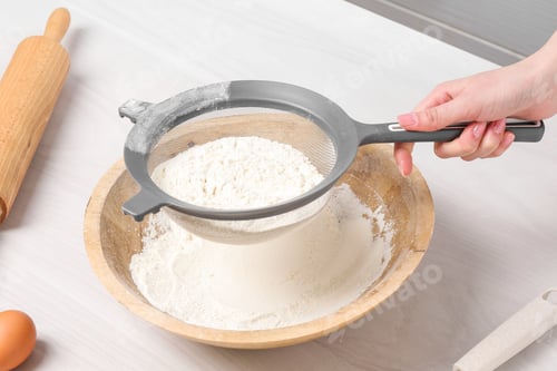 Preview: Woman sieving flour into bowl at white wooden table in kitchen, closeup