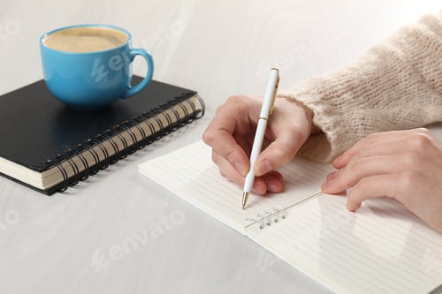 Preview: Woman writing in notebook near cup of refreshing coffee at light grey wooden table, closeup