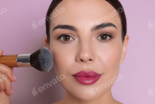 Preview: Beautiful woman applying makeup with brush on lilac color background, closeup