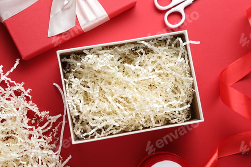 Preview: Gift box with shredded paper filler, ribbon and scissors on red background, flat lay