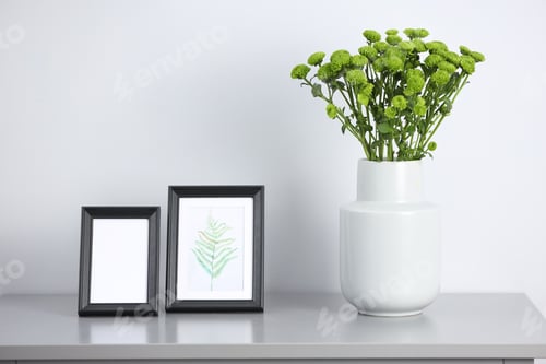 Preview: Green chrysanthemums in vase and picture frames on light grey table, closeup