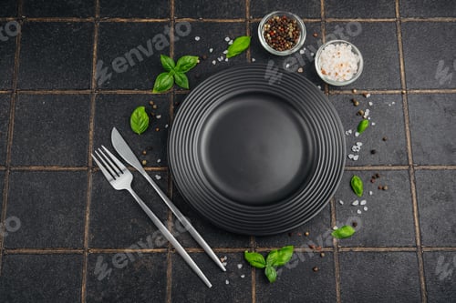 Preview: A black plate with fork and knife and basil on a dark background