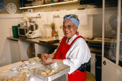 Preview: Happy Senior Black Woman Chef Smiling While Preparing Small Pastry Pies In A Professional Kitchen