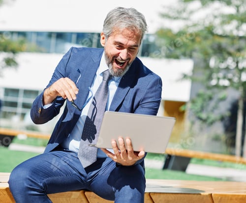 Preview: Successful Senior Businessman Laughing While Looking at Laptop Outdoors