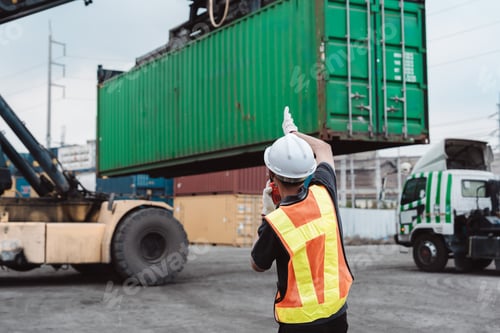 Preview: Logistics worker directing reach stacker lifting green container. Male operator using walkie-talkie