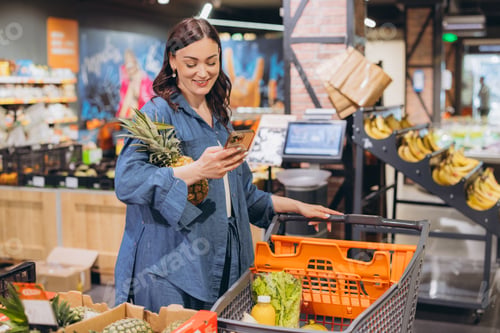 Preview: Woman standing in a grocery store aisle looking at her smartphone while shopping for fresh produce a