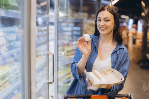 Preview: Young woman shopping for groceries, holding an egg carton and checking an egg in a supermarket food