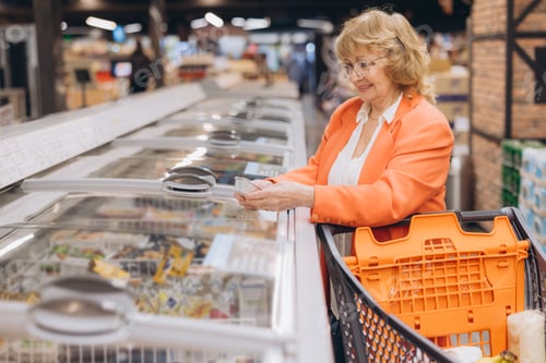 Preview: Senior woman smiling, shopping for frozen food in supermarket aisle, checking product at freezer che