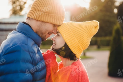 Preview: Young couple in love stand face to face outdoors at sunset, wear knitted hats and warm sweaters, sha