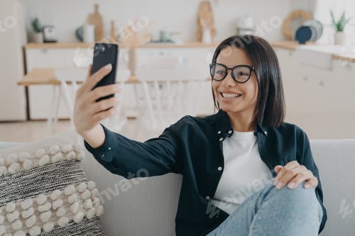 Preview: Smiling female in glasses holding phone, talks by video call, takes selfie photo, sitting on couch