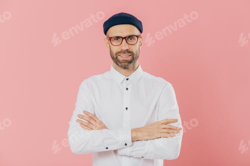 Preview: Indoor shot of self confident unshaven man looks happily at camera, keeps arms folded, wears headgea