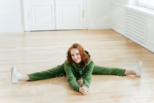 Preview: Horizontal shot of positive redhaired woman does pilates exercises, drssed in green tracksuit, has n
