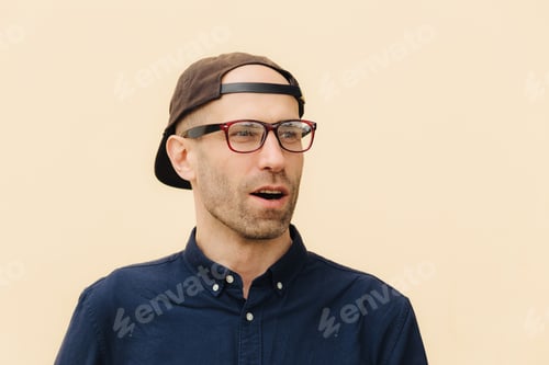 Preview: Surprised man wears backwards cap and glasses looks aside against beige wall