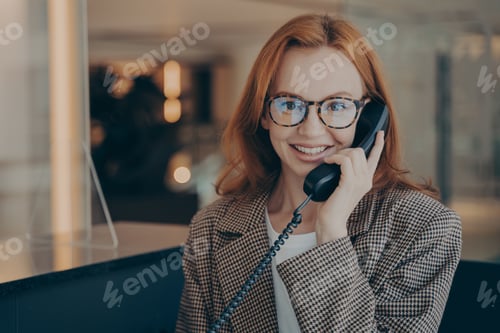Preview: Female office worker in spectacles talking on landline phone with co-worker and smiling at camera