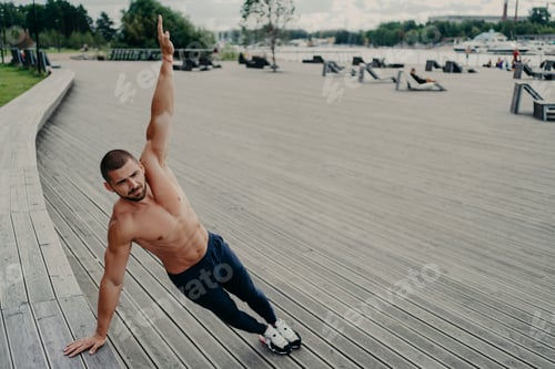 Preview: Self determined muscular man dressed in sport clothes stands in side plank, keeps arm raised, has wo