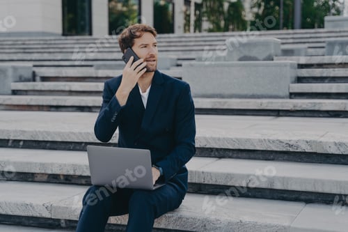 Preview: Professional Man Talking on Smartphone with Laptop Sitting on City Steps Outside Modern Office Build