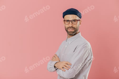 Preview: Studio shot of self assured bearded man with blue eyes, wears stylish hat and white shirt, keeps arm