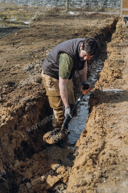 Preview: A man digs a drainage trench on his countryside property, working outdoors with a shovel