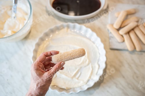 Preview: Woman holding ladyfinger biscuit over tiramisu cream preparation top view