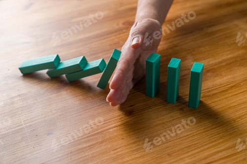 Preview: Hand stopping falling domino chain on wooden table risk control concept
