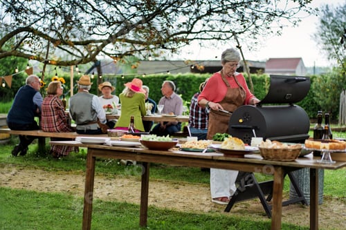 Preview: Senior woman grilling at garden barbecue while friends celebrate outdoors
