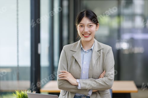Preview: Young asian businesswoman smiling, standing confidently with arms crossed in a modern office, portra