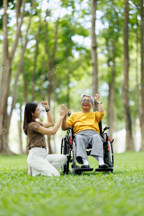 Preview: Asian caregiver providing joyful support to senior patient in wheelchair, sharing high five moment i