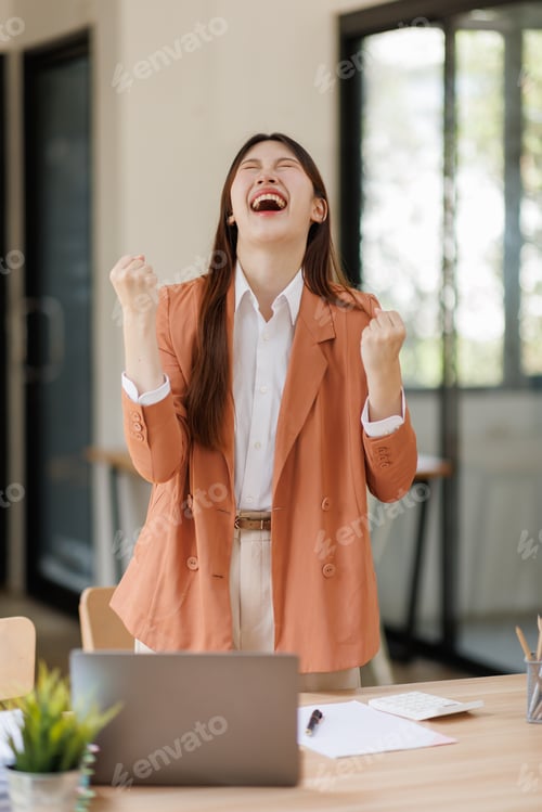 Preview: Asian businesswoman celebrating success with clenched fists in office