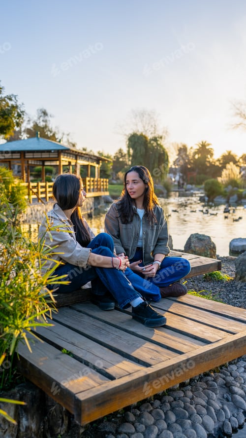 Preview: Two women relaxing on a wooden platform by a lake, enjoying a peaceful conversation at golden hour