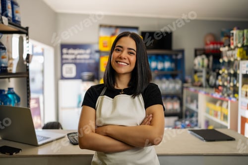 Preview: Young woman entrepreneur wearing an apron and smiling, crossing arms in a retail store