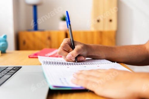 Preview: Unrecognizable young student girl writing notes while studying on laptop at home