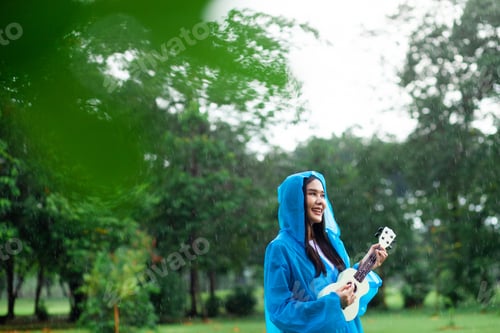 Preview: Beautiful Asian woman holding a ukulele feeling happy in the rain in the park. Concept of playing mu
