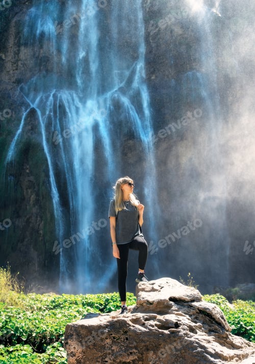 Preview: Young woman standing on a rock near big waterfall at sunset