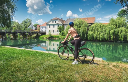Preview: Female cyclist with gravel bike standing on green grass and river