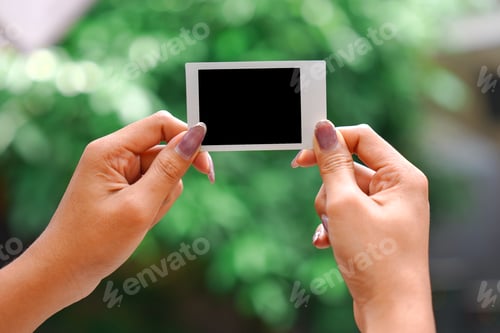 Preview: Close up of person's hand holding polaroid photo frame for mockup, outdoor nature background.