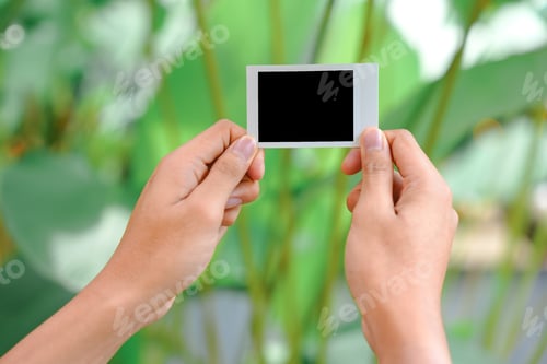 Preview: Close up of person's hand holding polaroid photo frame for mockup, outdoor nature background.