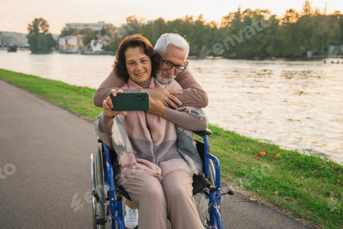 Preview: Senior couple woman in wheelchair caregiver old man making selfie enjoying walking. Elderly family m